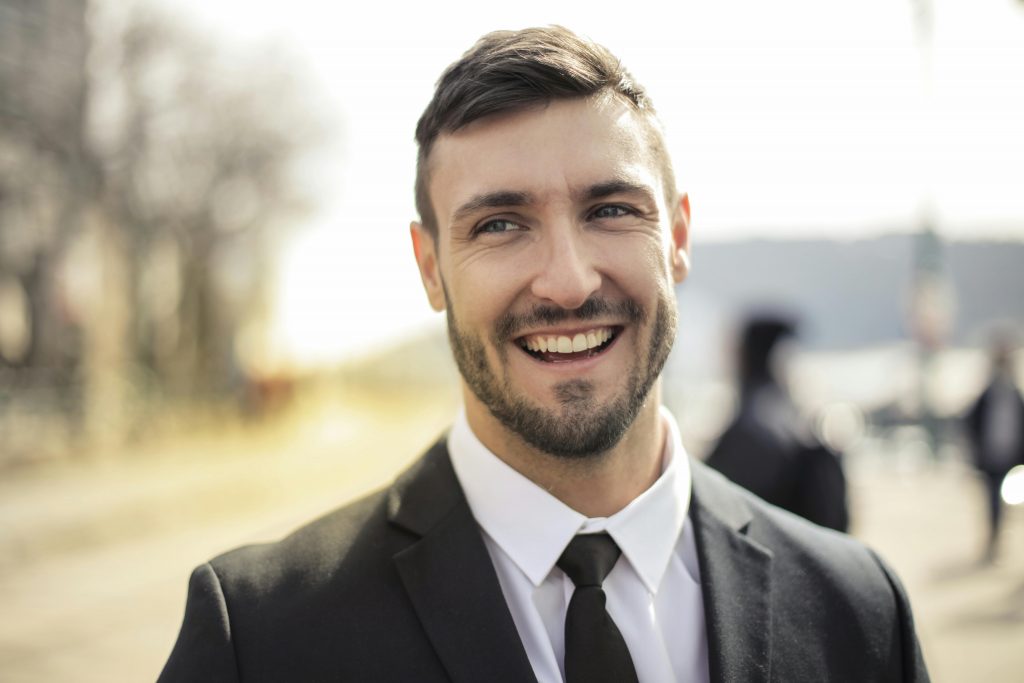 A confident businessman smiling in a black suit with an outdoor background.